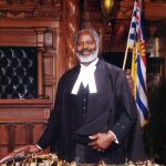 man in legislation robes standing behind desk , speakers mace on desk