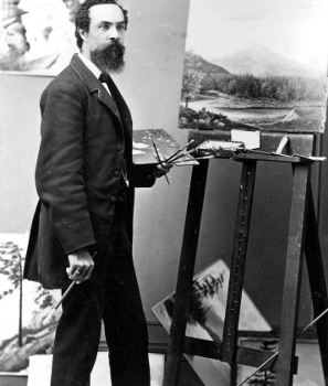 young bearded man standing at easel with paint brushes and palette in left hand and 1 brush in right hand