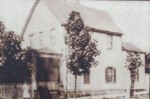 sepia toned taken from side angle of 2 story shingled and gabled house with porch, fence and trees at front house