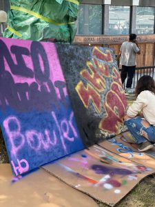 outside in park setting, woman crouching on right side/front facing 2 large graffiti works on plywood, applying finishing touches bottom right corner