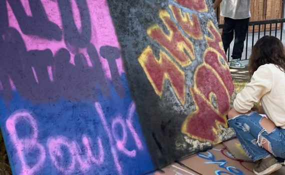 outside in park setting, woman crouching on right side/front facing 2 large graffiti works on plywood, applying finishing touches bottom right corner