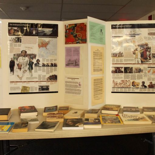 table with numerous books, magazines, brochures, free-standing poster at back with images, maps, text