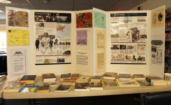 table with numerous books, magazines, brochures, free-standing poster at back with images, maps, text