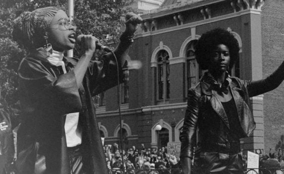 b&w public square 2 women on platform with raised fist 1 speaking at mic crowd in background