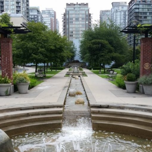 Park setting with concrete waterway running the length of the park, paved walkway, seating, greenspace on either side