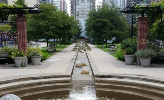 Park setting with concrete waterway running the length of the park, paved walkway, seating, greenspace on either side