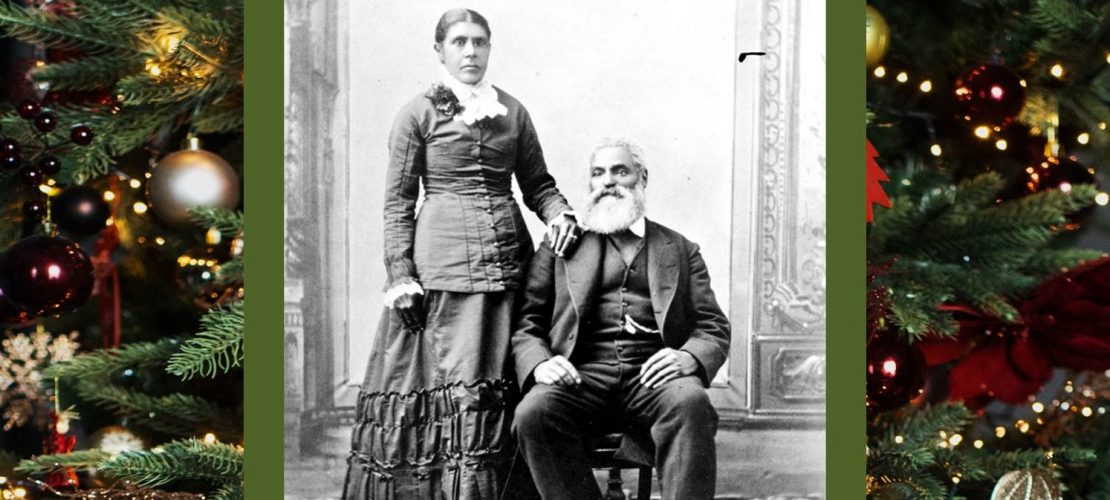 Black and white professional studio portrait woman standing, man seated. Nancy has dark hair parted in the centre, pulled back into a bun. Her left hand rests on Charles shoulder, seated, with his left foot slightly forward wearing a dark suit and vest, watch chain is visible from his vest pocket. Charles hair is white; he has a moustache and full beard. For this story, the image has a Christmas greenery border.