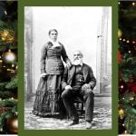 Black and white professional studio portrait woman standing, man seated. Nancy has dark hair parted in the centre, pulled back into a bun. Her left hand rests on Charles shoulder, seated, with his left foot slightly forward wearing a dark suit and vest, watch chain is visible from his vest pocket. Charles hair is white; he has a moustache and full beard. For this story, the image has a Christmas greenery border.