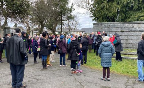 Group of about 50 people, women, men and youth, wearing coats/hats gathered around gravesites at Ross Bay Cemetery, speaker stands in front of one of the grave markers, grass underfoot, trees in background