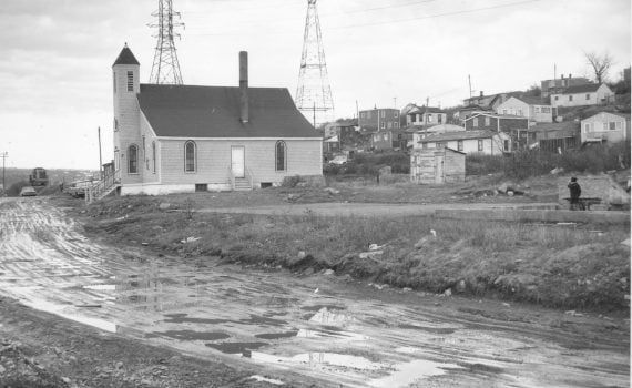 b&w side view of church prominent on right left side, dirt road, houses on slope on the right side (behind the church)