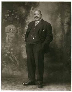 Full length portrait of black men, body slightly turned facing right, looking straight ahead, dressed in formal black suit, hands in pockets