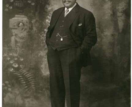 Full length portrait of black men, body slightly turned facing right, looking straight ahead, dressed in formal black suit, hands in pockets