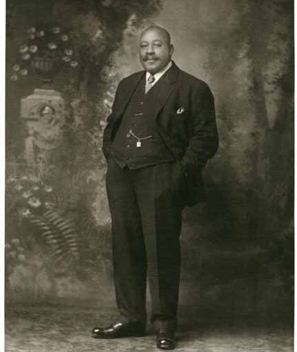 Full length portrait of black men, body slightly turned facing right, looking straight ahead, dressed in formal black suit, hands in pockets