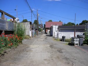 colour photo dirt road alley with housing, structures on either side