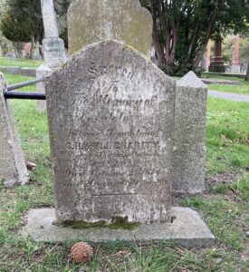 In cemetery, dark tarnished marble tablet with curved top on mossy concrete base