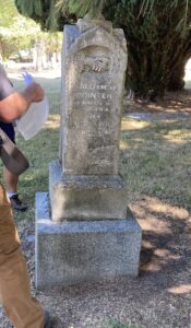 In cemetery, off-centre and leaning, stained, tarnished marble column on double concrete base with visible moss at the base