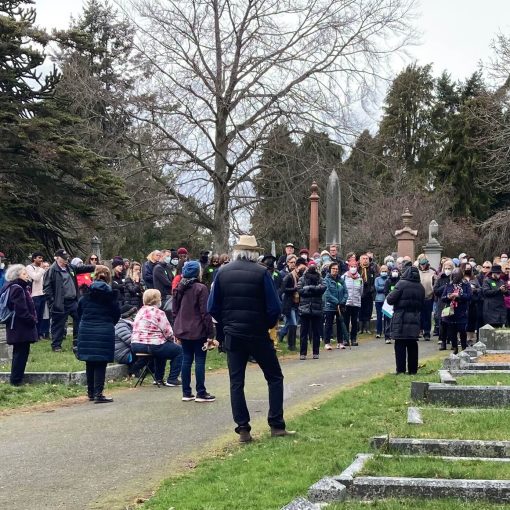 A large crowd of people gathered at the cemetery listen to a speaker, grave markers in the background and foreground