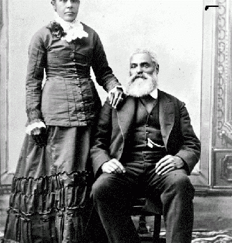 b&w professional studio portrait woman standing, man seated. Nancy's hair parted in the centre, pulled back into a bun. Her right hand rests on Charles shoulder. Charles, seated to her right, with his right foot slightly forward wearing a dark suit and vest, his watch chain is visible from his vest pocket. Charles hair is white; he has a moustache and full beard.