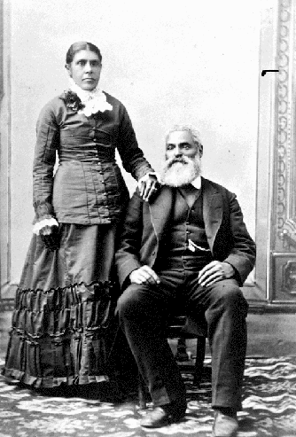 b&w professional studio portrait woman standing, man seated. Nancy's hair parted in the centre, pulled back into a bun. Her right hand rests on Charles shoulder. Charles, seated to her right, with his right foot slightly forward wearing a dark suit and vest, his watch chain is visible from his vest pocket. Charles hair is white; he has a moustache and full beard.