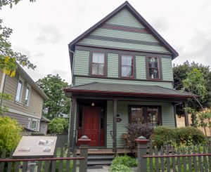 2 story front-gabled house, verandah, gray with burgundy trim, fenced garden