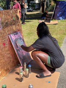 outside in park setting woman crouching working on graffiti piece on plywood, spray paint cans