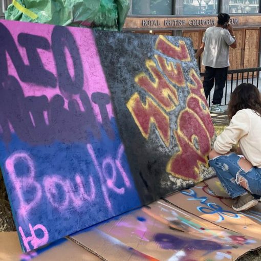 outside in park setting, woman crouching on right side/front facing 2 large graffiti works on plywood, applying finishing touches bottom right corner