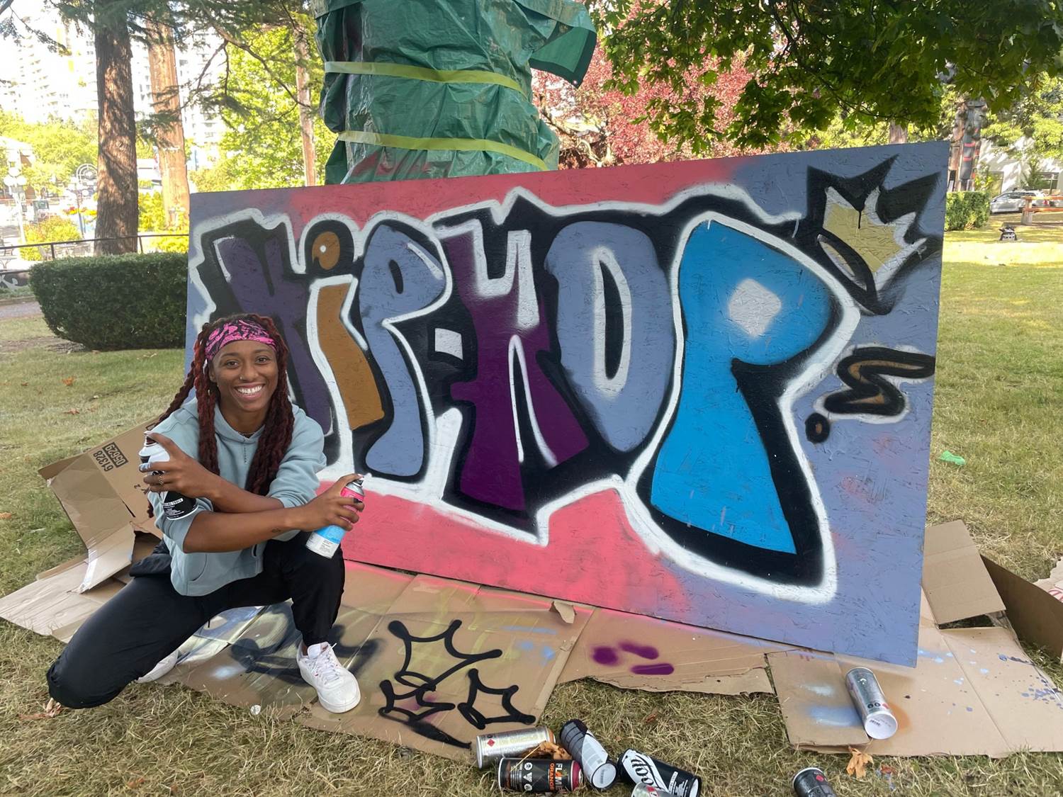 Woman crouching front left side of oversized completed graffiti work “Hip-Hop”; cans of spray paint in foreground