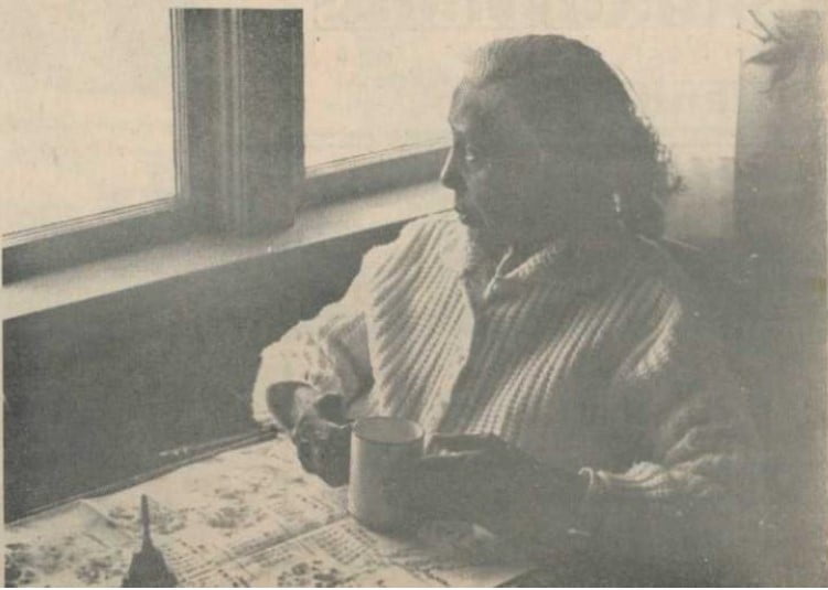 sepia photo, senior woman seated at table looking out a window, hands cupped around a mug placed on a newspaper open on the table