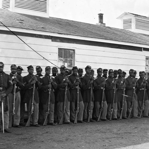 Black soldiers with their Black leader, lined up in 2 rows with rifles at their side, in front of a barracks