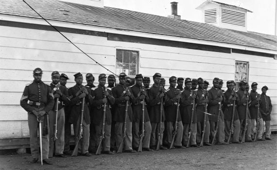 Black soldiers with their Black leader, lined up in 2 rows with rifles at their side, in front of a barracks