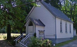 church with white clapboard siding, grey roof in treed setting. front steps to entryway