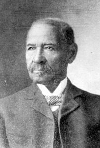 Portrait. Black male age 50, dressed semi-formally wearing jacket, vest, winged-collar white shirt with a striped pattern bow-tie