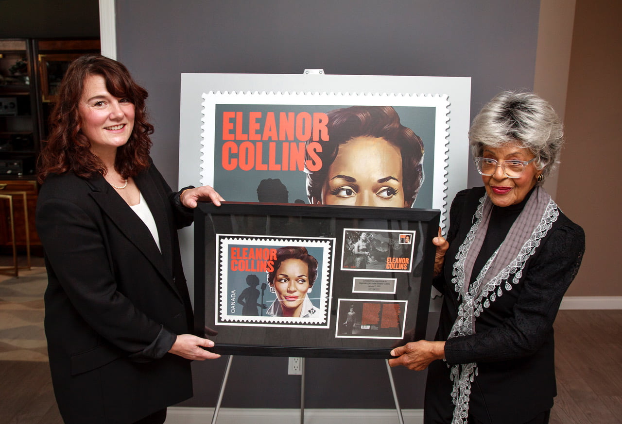 eleanor collins on right canada post female rep on left- standing holding commemorative stamp poster