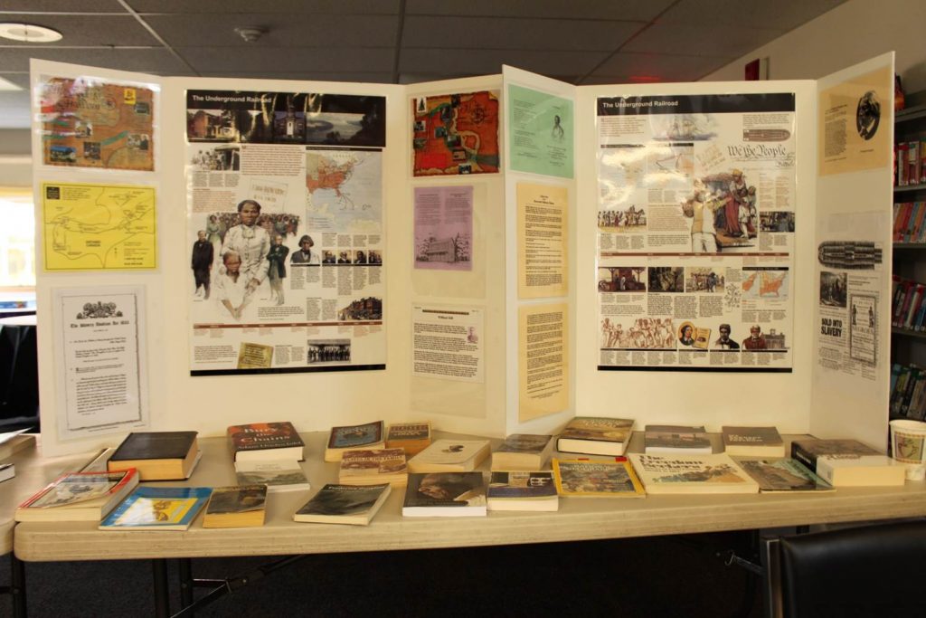 table with numerous books, magazines, brochures, free-standing poster at back with images, maps, text