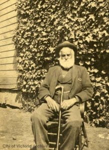 Sepia toned photo, man in senior years sitting in a chair with a cane leaning against his leg. In the background is a house and ivy hedge