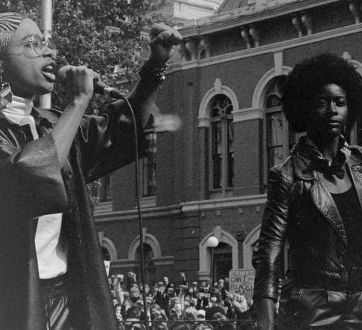 b&w public square 2 women on platform with raised fist 1 speaking at mic crowd in background