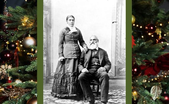 Black and white professional studio portrait woman standing, man seated. Nancy has dark hair parted in the centre, pulled back into a bun. Her left hand rests on Charles shoulder, seated, with his left foot slightly forward wearing a dark suit and vest, watch chain is visible from his vest pocket. Charles hair is white; he has a moustache and full beard. For this story, the image has a Christmas greenery border.