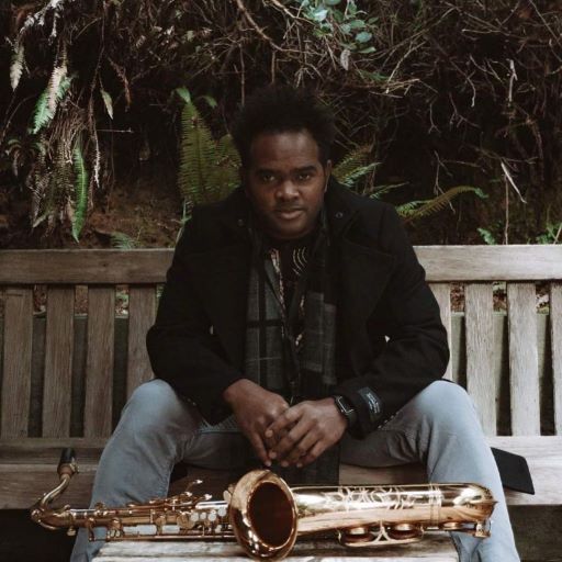 black man sitting on weathered bench looking directly at the camera, bent slightly forward, shrubbery in the background, foreground is saxophone on its side resting on low weathered table