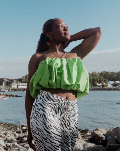 black women, face uplifted angled away from the camera, left arm bent with hand at side of head, wearing lime-green blouse-like halter top and white and dark patterned hip-hugger pants, at beach with buildings in the far background