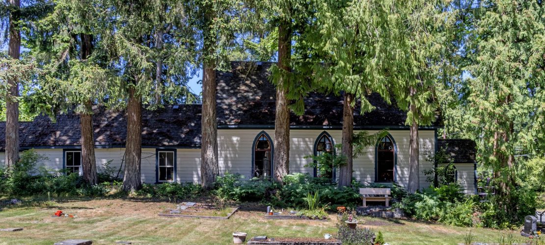 foreground is grassy cemetery with various grave markers, background is very tall trees, shrubbery with a side view of the Shady Creek Church with white clapboard siding, lancet-style windows along the side of the church