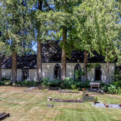 foreground is grassy cemetery with various grave markers, background is very tall trees, shrubbery with a side view of the Shady Creek Church with white clapboard siding, lancet-style windows along the side of the church