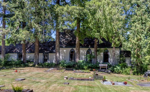 foreground is grassy cemetery with various grave markers, background is very tall trees, shrubbery with a side view of the Shady Creek Church with white clapboard siding, lancet-style windows along the side of the church
