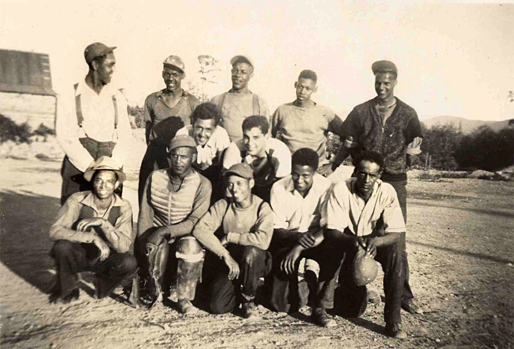 Sepia tone photo. 12 teenagers and young adults in three irregular rows, standing, crouching or kneeling. Dressed casually, smiling, wearing a variety of clothing, hats and caps.