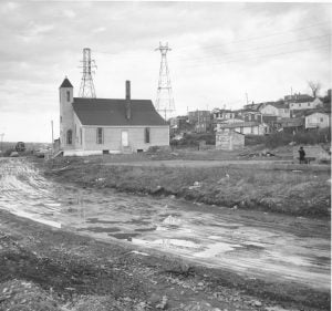 b&w side view of church prominent on right left side, dirt road, houses on slope on the right side (behind the church)