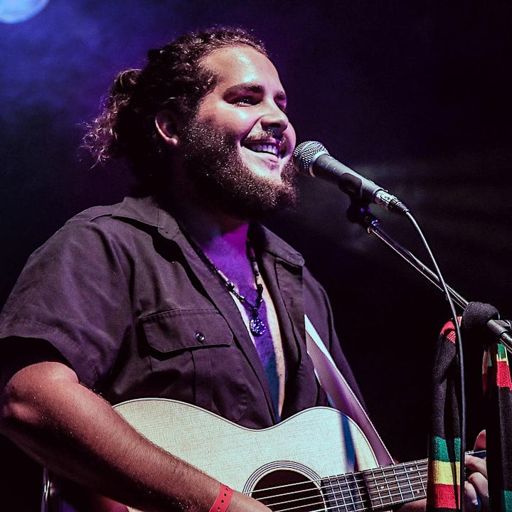 upper torso man, smiling, hair in ponytail on stage at mic playing guitar