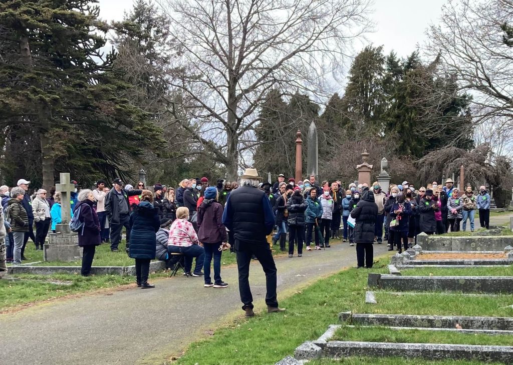 A large crowd of people gathered at the cemetery listen to a speaker, grave markers in the background and foreground
